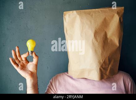 Mann mit einer Papiertüte auf dem Kopf, der die Glühbirne in der Hand hält. Ideenkonzept Stockfoto