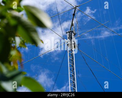 Eine hohe HAM-Radioantenne vor einem blau bewölkten Himmel hinter grünen Blättern. Stockfoto