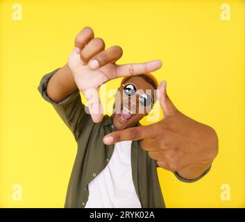 Ein junger kreativer Mann auf gelbem Hintergrund wird gesehen, wie er aus seinen Fingern einen Rahmen für die Komposition bildet, der seine künstlerischen und imaginativen Fähigkeiten zeigt Stockfoto