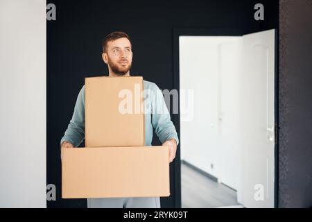 Junger lächelnder Mann zieht in eine neue Wohnung mit Pappkartons Stockfoto