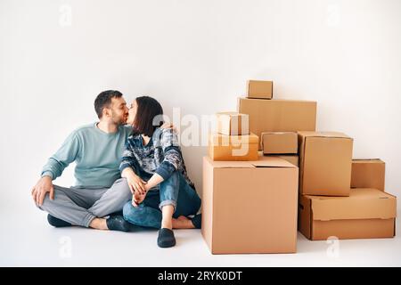 Junge, liebevolle Paare küssen sich auf dem Boden in ihrer neuen Wohnung am Umzugstag, umgeben von Pappschachteln Stockfoto