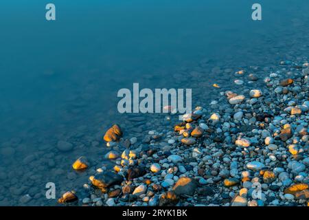 Bunte Kieselsteine am blauen Flussufer, fotografiert in den letzten Minuten des goldenen Sonnenuntergangs, färben einige der Steine in Orange Stockfoto