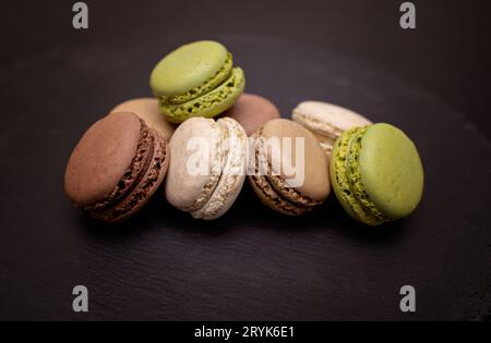 Macarons in Beige-, Braun-, Pistazien- und Cremetönen auf schwarzem Schieferhintergrund. Nahaufnahme in verschiedenen Positionen. Selektiver Fokus. Stockfoto