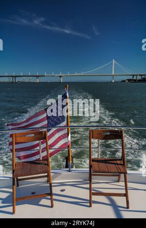 Stühle auf dem Deck einer Yacht, die die Bucht von San Francisco überquert, mit amerikanischer Flagge und Bay Bridge im Hintergrund Stockfoto