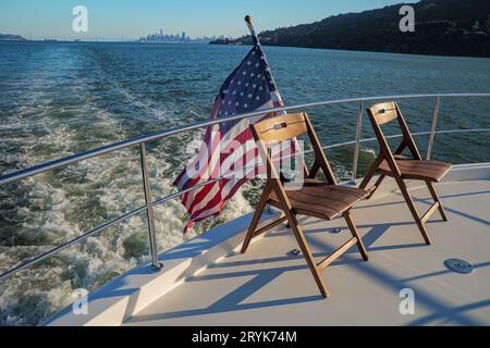 Stühle auf dem Deck einer Yacht, die die Bucht von San Francisco überquert, mit amerikanischer Flagge und Stadt im Hintergrund Stockfoto