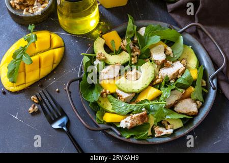 Gesunder Diätsalat mit Rucola, Mango, Avocado, Hühnergrill, Walnüssen und Vinaigrette-Dressing auf einer dunklen Steinplatte. Stockfoto