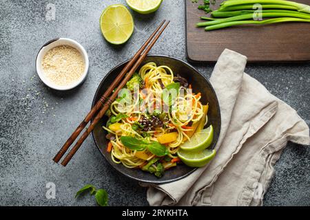 Asiatische vegetarische Nudeln mit Gemüse und Limette in schwarzer rustikaler Keramikschale, Holzstäbchen, Schneidebrett mit gehacktem Gr Stockfoto