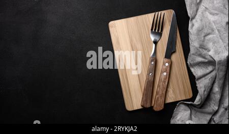 Gabel und Messer mit Holzgriff und Schneidbrett auf schwarzem Hintergrund, Draufsicht Stockfoto