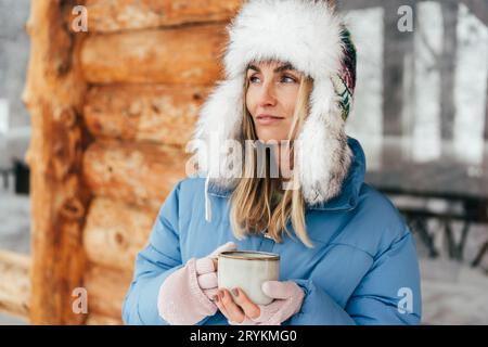 Porträt einer kaukasischen Frau in einem Winterhut mit Ohrenklappen mit einem Becher heißen Getränks. Stockfoto