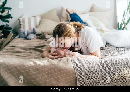 Eine zarte Mutter, die auf dem Bett liegt, umarmt und küsst das Baby. Authentisches Familienportrait. Mutterschaft. Stockfoto