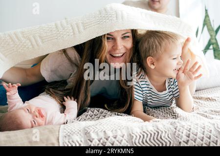 Fröhliche Mama spielt mit den Kindern auf dem Bett und bedeckt die Kinder und sich mit einer Decke. Spaß Familie spielerische Zeit. Stockfoto
