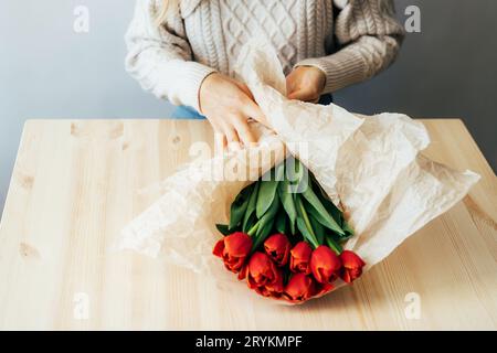 Nahaufnahme der weiblichen Hände, die rote Tulpen in Papier verpackt haben. Tageslicht am Arbeitsplatz eines Floristen in Frühlingsstimmung. Stockfoto