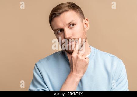 Nachdenklicher, rätselhafter, wehmütiger Mann, der das Kinn auf beigem Hintergrund berührt. Stockfoto