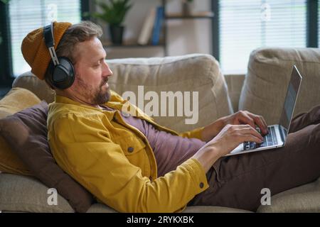 Junger Mann in lässiger Kleidung, der auf dem Sofa sitzt und Online-Spiel auf dem Laptop spielt. Er konzentriert sich voll auf das Spiel und trägt Kopf Stockfoto