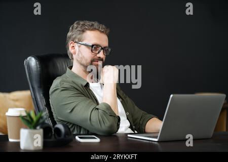 Weiser, kaukasischer Mann im Portrait, sitzt am Schreibtisch im Heimbüro. Er wirkt nachdenklich, während er am Computer arbeitet. Sens Stockfoto