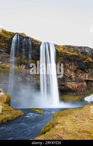 Malerische isländische Kaskade umgeben von hohen Bergen und wunderbarem Grün. Der Wasserfall Seljalandsfoss rauscht über einen Rand mit eiskaltem Wasser und großen Steinen, nordische Ziele. Stockfoto