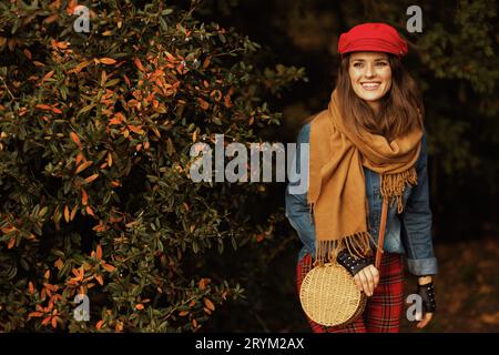 Hallo Herbst. Glückliche, trendige 40-jährige Frau im Jeanshemd und roten Hut mit Schal, Handschuhen und Tasche im Stadtpark. Stockfoto