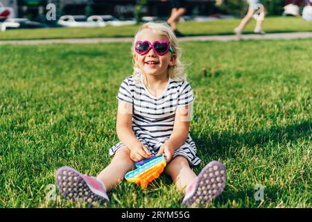 Ein kleines bezauberndes lächelndes, dreijähriges Mädchen sitzt mit einer Sonnenbrille auf dem Rasen und spielt mit einem Spielzeug. Stockfoto