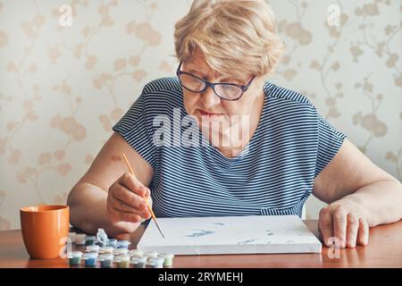 Attraktive alte Frau, die zu Hause nach Zahlen malte. Inspirierende Hobby- und Kunsttherapie. Selektiver Fokus Stockfoto