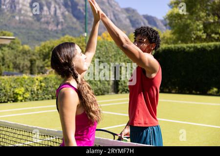 Glückliches, vielseitiges Paar mit Schlägern, High Fiving über dem Netz auf einem sonnigen Tennisplatz im Freien Stockfoto