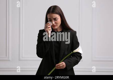 Traurige Frau mit Calla-Lilie-Blume, die nahe der weißen Wand trauert. Trauerzeremonie Stockfoto