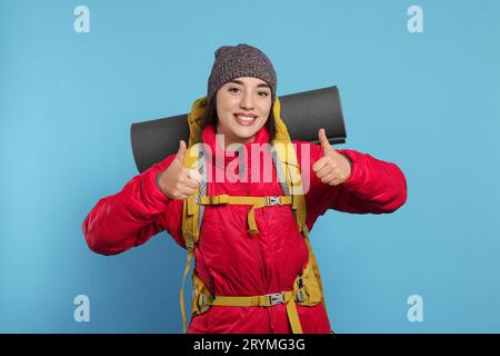 Lächelnde junge Frau mit Rucksack, der Daumen hoch auf hellblauem Hintergrund zeigt. Aktiver Tourismus Stockfoto