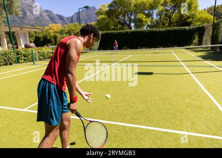 Ein birassistischer Mann mit einem Raketenball, der einem Spieler auf einem sonnigen Tennisplatz im Freien dient, Kopierbereich Stockfoto