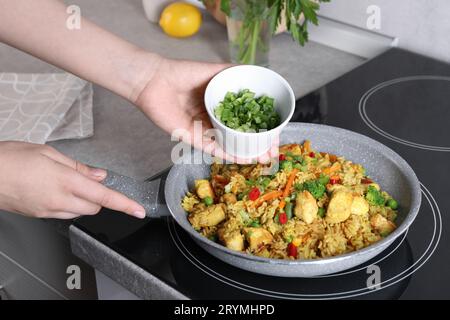 Frau fügt geschnittene grüne Zwiebeln zu Reis mit Fleisch und Gemüse in der Pfanne hinzu, Nahaufnahme Stockfoto