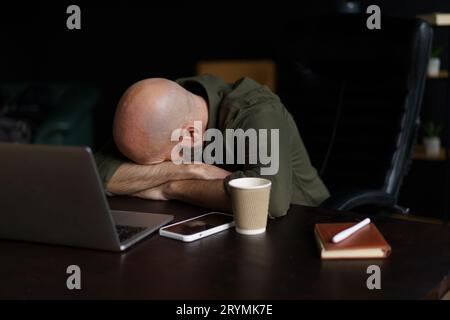 Müder Arbeiter mittleren Alters schläft mit dem Kopf auf dem Tisch in der Nähe des Laptops. Das Konzept altersbedingter Veränderungen wird dargestellt, wobei die Bedeutung von Imp Stockfoto