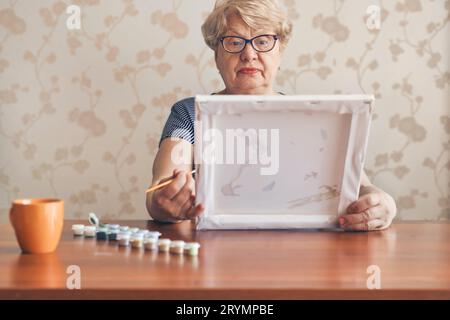 Attraktive alte Frau, die zu Hause nach Zahlen malte. Inspirierende Hobby- und Kunsttherapie. Selektiver Fokus Stockfoto