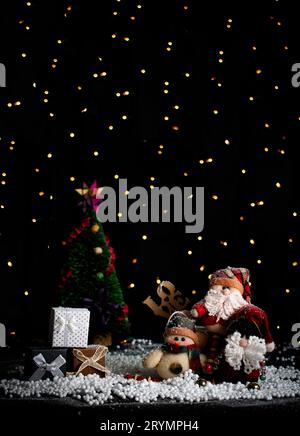 Weihnachtskarte mit 3 plüschigen Weihnachtsmännern, 3 Geschenkboxen und im Hintergrund einem Weihnachtsbaum vor schwarzem Hintergrund mit verschwommenem Christma Stockfoto