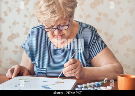 Attraktive alte Frau, die zu Hause nach Zahlen malte. Inspirierende Hobby- und Kunsttherapie. Selektiver Fokus Stockfoto