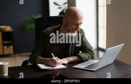 Lächelnder Mann im mittleren Alter, fokussiert und produktiv. Er saß drinnen am Schreibtisch, vertiefte die Arbeit. Mit Notizblock vorne, er fleißig Stockfoto