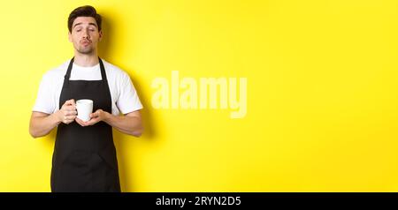 Mann in schwarzer Schürze, der eine Tasse Kaffee bringt und auf einen Kuss wartet, der auf gelbem Hintergrund steht Stockfoto