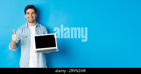 Ein lächelnder, gutaussehender Mann zeigt den Laptop-Bildschirm und den Daumen nach oben, wie ein Werbeangebot, empfahl die Website, steht vor blauem Hintergrund Stockfoto