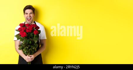 Lustiger junger Verkäufer in schwarzer Schürze, der Strauß Rosen hält, Blumenhändler lachend und über gelbem Hintergrund stehend Stockfoto