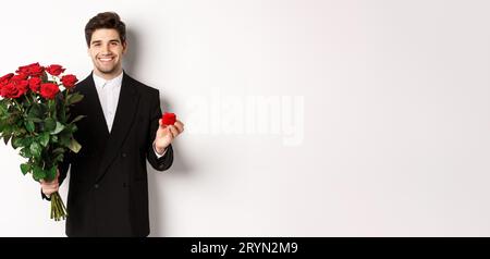 Bild von schönen Mann in schwarzem Anzug, hält Strauß von roten Rosen und einen Ring, einen Vorschlag, lächelnd zuversichtlich, stehend aga Stockfoto