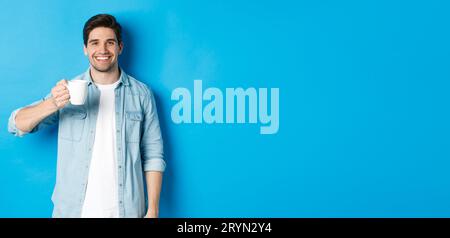 Lächelnder bärtiger Mann hält Becher und trinkt Kaffee, steht vor blauem Hintergrund Stockfoto