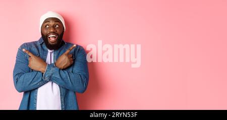 Aufgeregter junger afroamerikanischer Mann, der seitwärts zeigt, zwei Entscheidungen zeigt und eine Entscheidung trifft, glücklich lächelt, über rosa steht Stockfoto