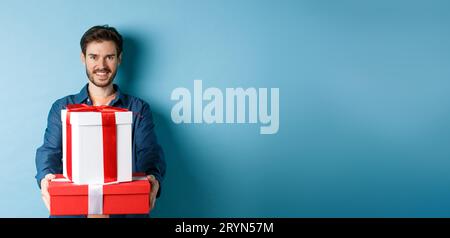 Alles gute zum Valentinstag. Hübscher Mann, der Freundinnen Geschenke gibt, Geschenkboxen hält und lächelt, auf blauem Hintergrund stehend Stockfoto