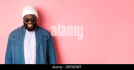 Cooler afroamerikanischer Hipster-Mann in Beanie und Sonnenbrille, lächelnd und in die Kamera schauend, selbstbewusst über Rosa Stockfoto