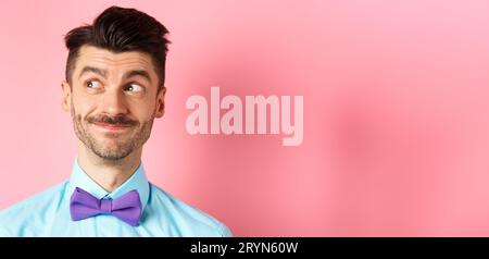 Nahaufnahme eines gutaussehenden lächelnden Kerls mit Schnurrbart, der links auf das Logo mit dem fröhlichen Gesicht blickt und auf einem rosa Hintergrund steht Stockfoto