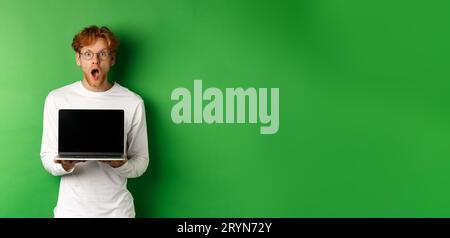 Überraschter Rothaariger Mann in Brille, zeigt einen leeren Laptop-Bildschirm, zeigt Online-Promo und Kieferkiefer, starrt die Kamera staunend, grün Stockfoto