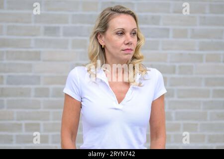 Oberkörperporträt einer blonden Frau, die nachdenklich auf die Seite vor einer weißen Ziegelwand schaut Stockfoto