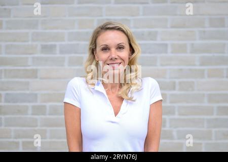 Oberkörperporträt einer attraktiven blonden Frau, die vor einer weißen Ziegelwand auf die Kamera lächelt Stockfoto