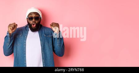 Der afroamerikaner mit Sonnenbrille und Beanie gewinnt und freut sich, lässt die Faust triumphieren, starrt in die Kamera und Stockfoto