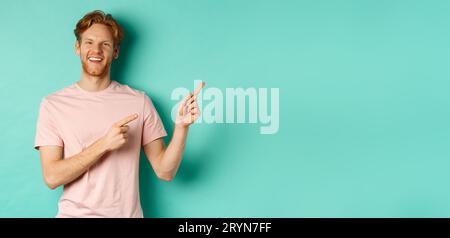 Hübsches männliches Model mit rotem, unordentlichem Haar, das Werbung auf dem Kopierraum zeigt, auf die rechte obere Ecke zeigt und glücklich lächelt, s Stockfoto