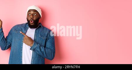 Beeindruckender schwarzer Hipster-Mann in Mütze und Denim-Hemd, der auf das Logo in der oberen linken Ecke zeigt, staunend und stehend starrt Stockfoto