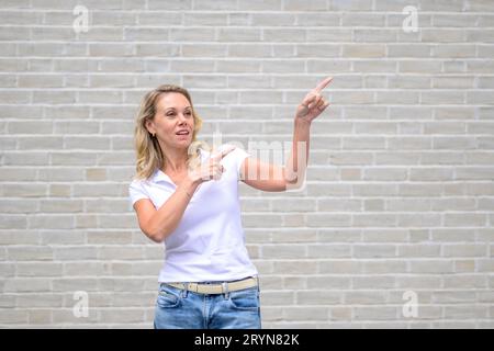 Nahaufnahme eines Porträts einer attraktiven, freundlichen Frau, die auf die rechte Seite und nach oben vor eine weiße Backsteinmauer zeigt Stockfoto