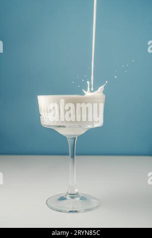 Milch gießt in ein wunderschönes Kristallglas auf blauem Hintergrund und spritzt herum. Stockfoto